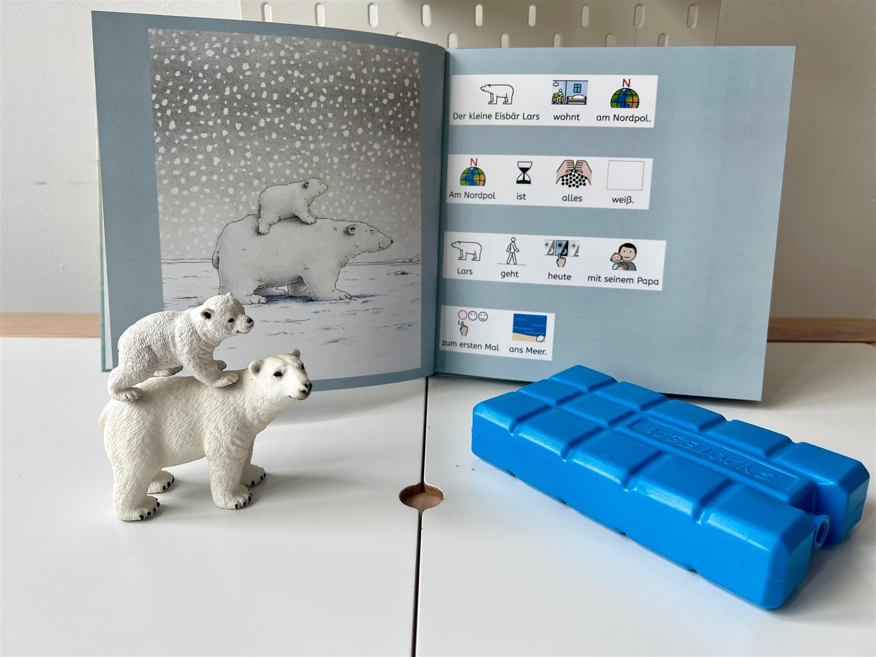 Aufgeschlagenes IncluBook: links eine Bilderbuchseite mit Eisbären, rechts kurze Sätze mit METACOM-Symbolen. Davor stehen zwei Eisbär-Figuren und ein blaues Instrument.